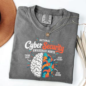 May include: A charcoal gray short-sleeve t-shirt featuring a graphic of a split brain. The left side is white, and the right side is multicolored. Text reads: National Cyber Security Awareness Month, Think Safe, Stay Alert, and Stay Secure.