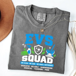May include: A charcoal gray t-shirt featuring the blue text EVS SQUAD and a graphic of a spray bottle, a shield, and a mop. Below the graphic, the text reads WE CLEAN WE CARE WE MAKE A DIFFERENCE, followed by DEDICATED, RELIABLE, HARDWORKING, ESSENTIAL, TEAMWORK, PRIDE.