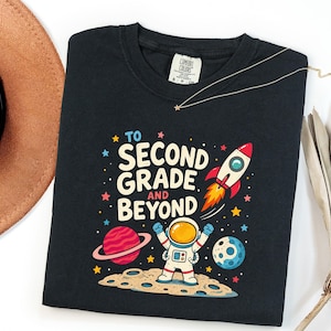 May include: A black crewneck t-shirt featuring a colorful space-themed graphic with an astronaut standing on a moon surface, a rocket ship, planets, and stars. The text reads TO SECOND GRADE AND BEYOND in white and red lettering on the front.