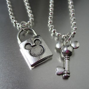 May include: A silver chain necklace with a padlock pendant featuring a Mickey Mouse silhouette and a matching key pendant with a Mickey Mouse silhouette.