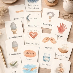 May include: A set of watercolour cards featuring phrases like "Thank You," "Bathroom," and "I Need Water," with accompanying illustrations. The cards are displayed on a wooden surface, with a coffee cup and a pen also visible.
