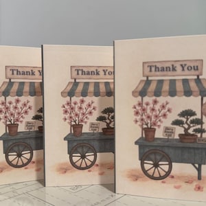May include: Three greeting cards with a watercolor illustration of a cart with cherry blossoms and bonsai trees. The cart has a striped awning and the words "Thank You" at the top.