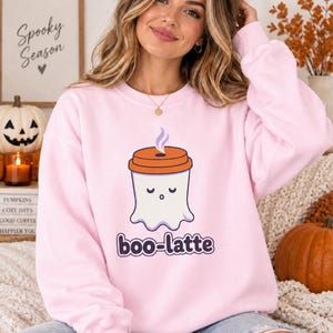 Könnte beinhalten: Ein hellrosa Sweatshirt mit Rundhalsausschnitt und einer Grafik eines weißen Geistes in Form eines Kaffeebechers mit orangefarbenem Deckel und einer Dampfwolke. Unter dem Geist steht der Text "boo-latte" in dunkelvioletten, runden, fetten Kleinbuchstaben.