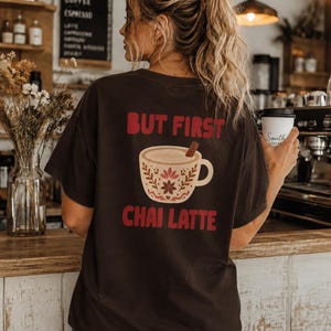 Könnte beinhalten: Ein dunkelbraunes Kurzarm-T-Shirt mit einem Grafikdruck auf der Rückseite. Das Design zeigt den roten Schriftzug "BUT FIRST" über einer beigen Tassenillustration mit Blumenmustern und einer Zimtstange, gefolgt vom roten Text "CHAI LATTE".