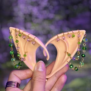 May include: A pair of pale, pointed ear cuffs made of a smooth, skin-toned material. Each cuff features gold-toned metal chains adorned with small, faceted green and light purple crystal beads that drape across the surface of the ear shape.
