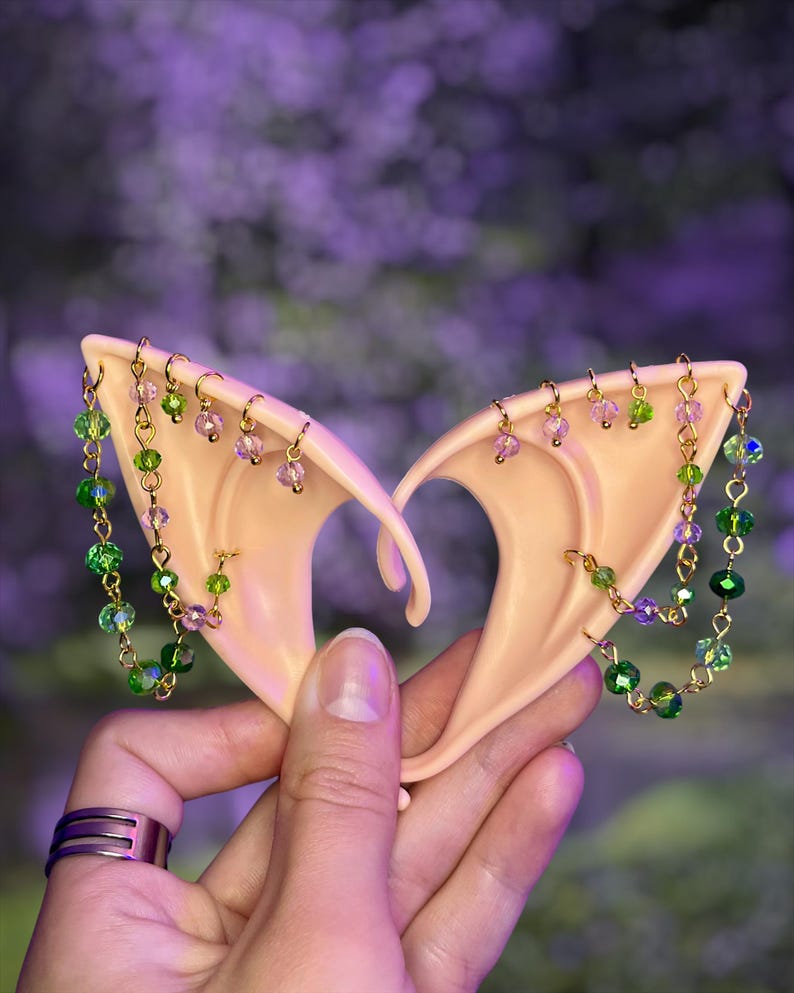 May include: A pair of pale, pointed ear cuffs made of a smooth, skin-toned material. Each cuff features gold-toned metal chains adorned with small, faceted green and light purple crystal beads that drape across the surface of the ear shape.