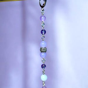 May include: A dangling earring with a gold-colored top and a chain of purple and blue beads. The beads vary in size and shade, with a decorative silver bead in the center. The background is a soft purple.