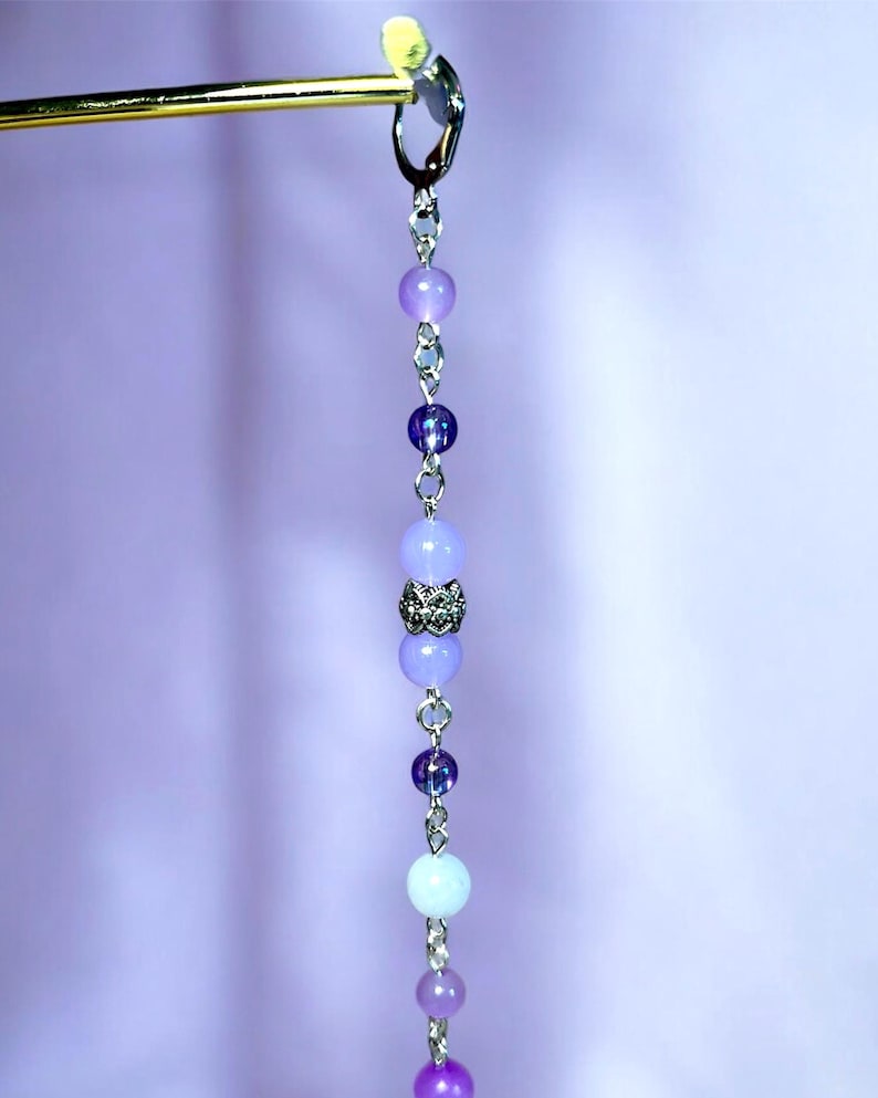 May include: A dangling earring with a gold-colored top and a chain of purple and blue beads. The beads vary in size and shade, with a decorative silver bead in the center. The background is a soft purple.
