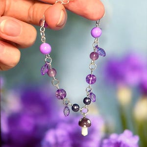 May include: A delicate silver chain bracelet featuring a whimsical design. The bracelet is adorned with various shades of purple beads, including round and faceted styles, along with small leaf-shaped charms. A miniature mushroom charm dangles from the center.