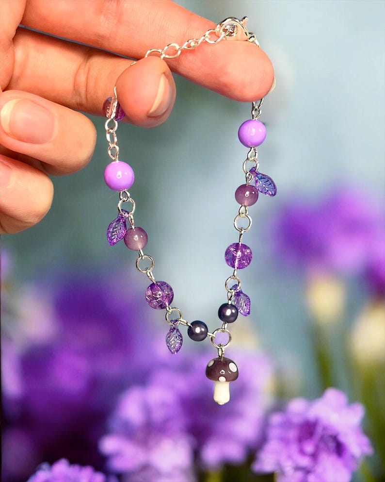 May include: A delicate silver chain bracelet featuring a whimsical design. The bracelet is adorned with various shades of purple beads, including round and faceted styles, along with small leaf-shaped charms. A miniature mushroom charm dangles from the center.