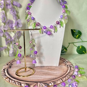 Op de afbeelding: Een sieradenset bestaande uit een ketting en bijpassende oorbellen, gemaakt met paarse en roze kralen, doorschijnende groene bladbedels en kleine paarse klokvormige bloemhangers aan zilverkleurige metalen kettingen en onderdelen.