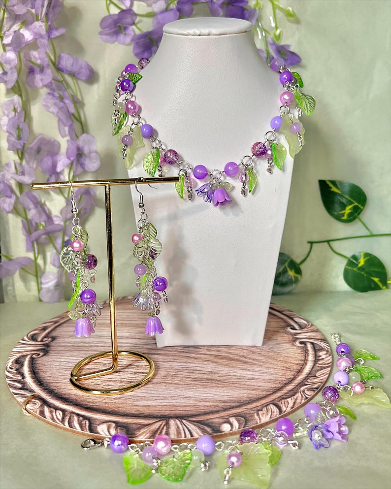 Op de afbeelding: Een sieradenset bestaande uit een ketting en bijpassende oorbellen, gemaakt met paarse en roze kralen, doorschijnende groene bladbedels en kleine paarse klokvormige bloemhangers aan zilverkleurige metalen kettingen en onderdelen.