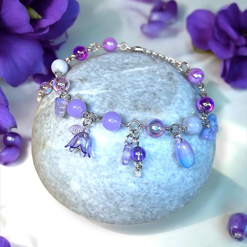 May include: A delicate bracelet with a silver chain and a variety of purple beads and charms. The bracelet features iridescent, lavender, and deep purple beads, along with flower and teardrop charms. The bracelet is displayed on a gray stone.