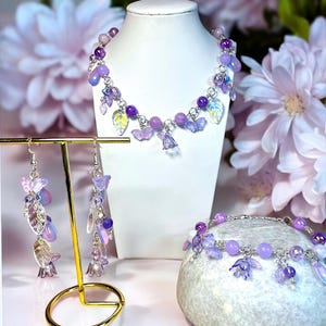 Puede incluir: Un conjunto de joyas que incluye un collar, una pulsera y pendientes. El conjunto incluye cuentas moradas, dijes de mariposa y colgantes en forma de hoja. Las joyas se exhiben en un busto blanco y un soporte dorado para pendientes.