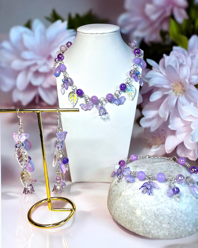 Puede incluir: Un conjunto de joyas que incluye un collar, una pulsera y pendientes. El conjunto incluye cuentas moradas, dijes de mariposa y colgantes en forma de hoja. Las joyas se exhiben en un busto blanco y un soporte dorado para pendientes.