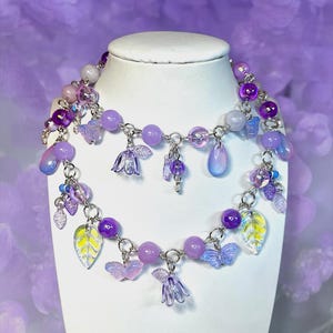 May include: A two-strand necklace displayed on a white bust. The necklace features a variety of purple beads, charms, and pendants. Charms include butterflies, leaves, and flower shapes. The background is a soft purple.