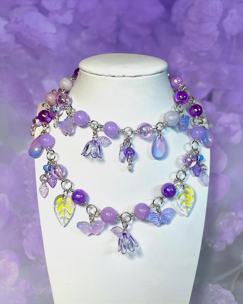 May include: A two-strand necklace displayed on a white bust. The necklace features a variety of purple beads, charms, and pendants. Charms include butterflies, leaves, and flower shapes. The background is a soft purple.