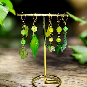 May include: A collection of six green and gold earrings displayed on a gold-colored earring stand. The earrings feature various leaf and bead designs in shades of green, with gold-colored hooks and chains. The stand is on a wooden surface.