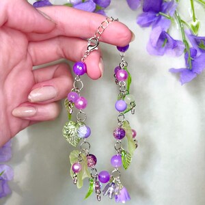Op de afbeelding: Een bedelarmband met een zilverkleurige ketting versierd met diverse paarse en groene kralen, doorschijnende bladvormige bedels en kleine klokvormige bloemhangers in tinten lichtpaars en helder glas, afgewerkt met een karabijnsluiting.
