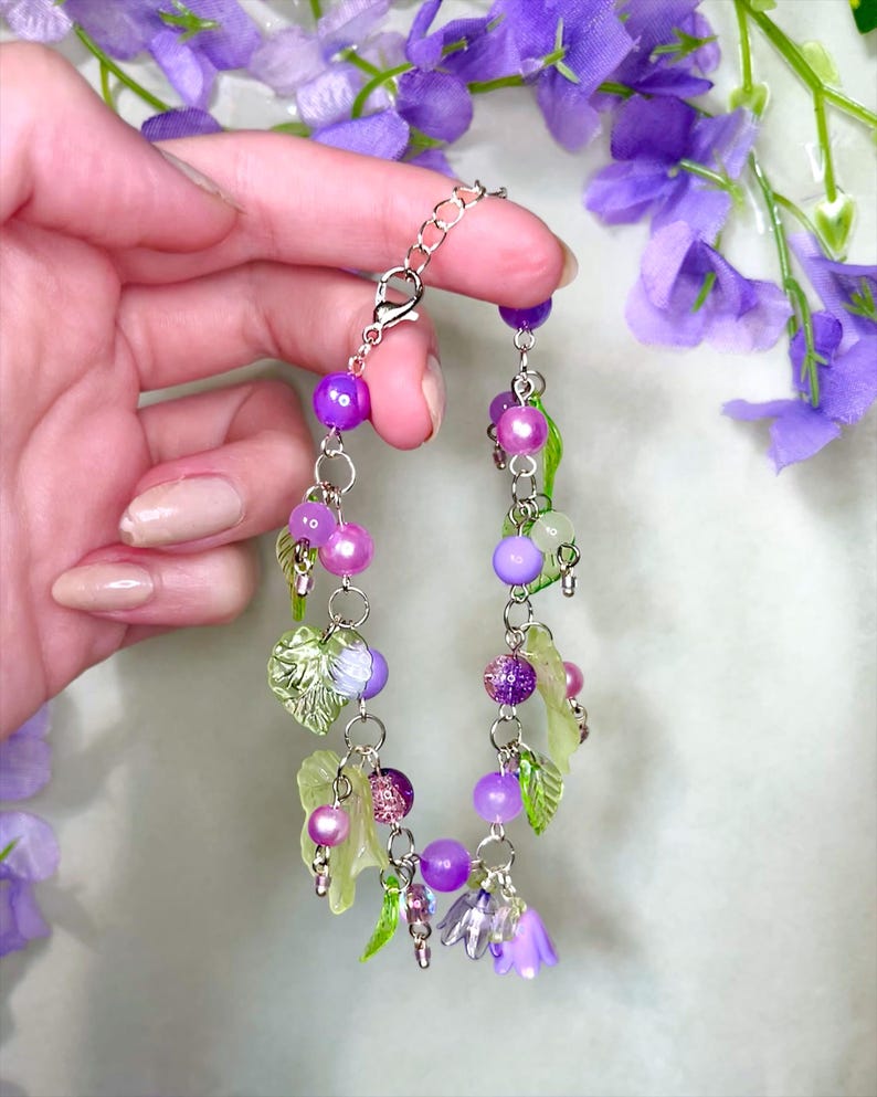 Op de afbeelding: Een bedelarmband met een zilverkleurige ketting versierd met diverse paarse en groene kralen, doorschijnende bladvormige bedels en kleine klokvormige bloemhangers in tinten lichtpaars en helder glas, afgewerkt met een karabijnsluiting.