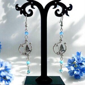 May include: Silver-tone dangle earrings featuring a butterfly design within a circular frame. The earrings have light blue beads and a small, dangling crystal. The earrings are displayed on a black stand, with blue flowers in the background.