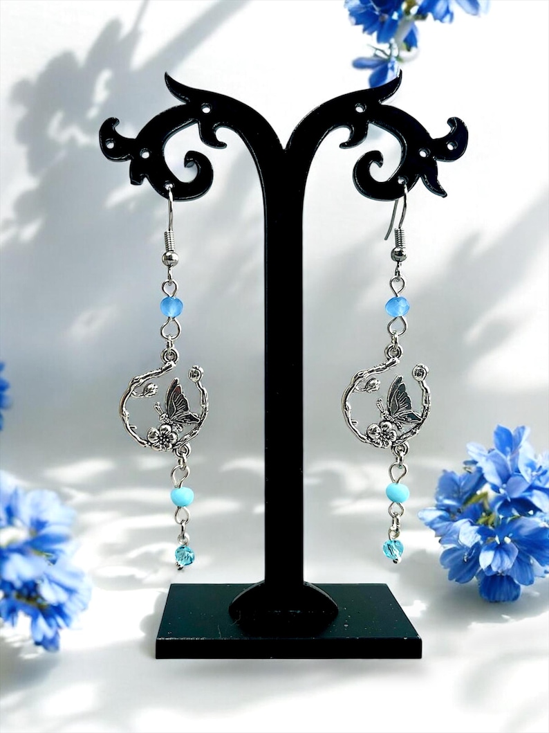 May include: Silver-tone dangle earrings featuring a butterfly design within a circular frame. The earrings have light blue beads and a small, dangling crystal. The earrings are displayed on a black stand, with blue flowers in the background.