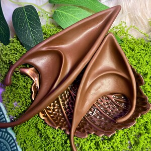 May include: A pair of pointed, brown, ear-shaped prosthetics with a smooth, curved design. These accessories feature a long, tapered tip and a contoured base, crafted to mimic the appearance of fantasy-style ears with a solid, matte finish.