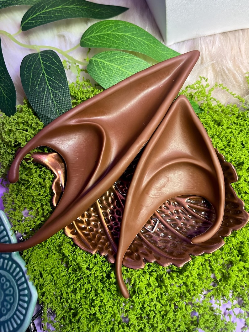 May include: A pair of pointed, brown, ear-shaped prosthetics with a smooth, curved design. These accessories feature a long, tapered tip and a contoured base, crafted to mimic the appearance of fantasy-style ears with a solid, matte finish.