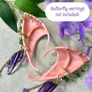 May include: A pair of peach-colored pointed ear cuffs featuring gold-tone chains adorned with small green and purple beads. Each cuff includes a dangling translucent purple wing-shaped charm. The text (butterfly earrings not included) is visible.