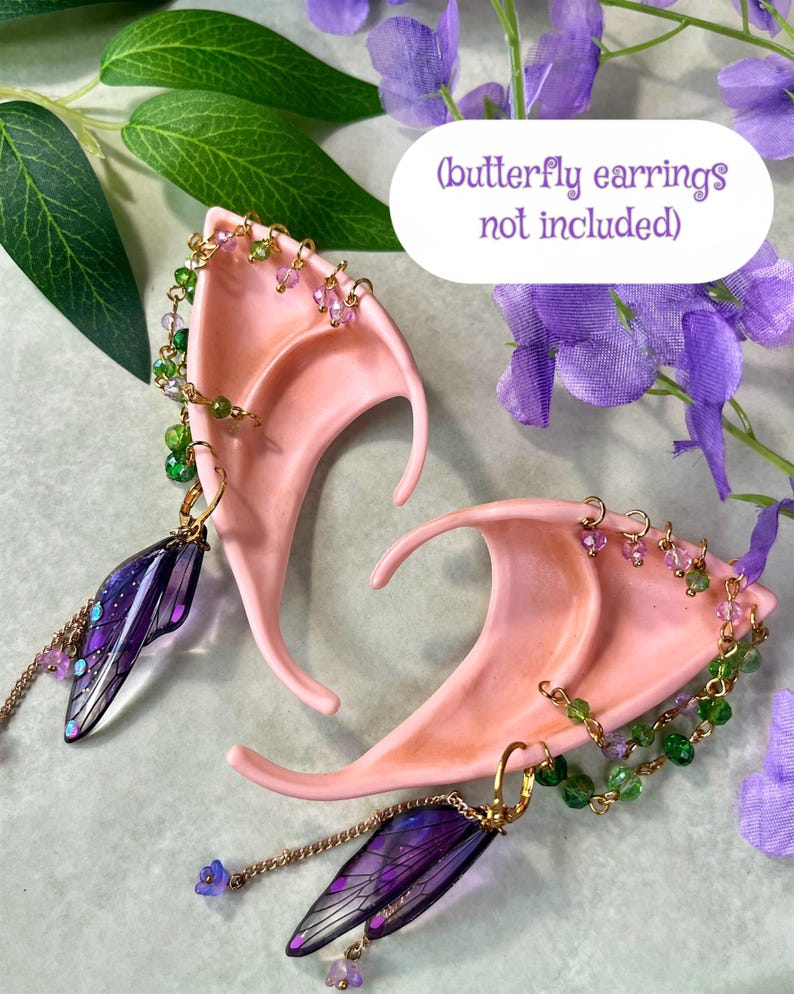 May include: A pair of peach-colored pointed ear cuffs featuring gold-tone chains adorned with small green and purple beads. Each cuff includes a dangling translucent purple wing-shaped charm. The text (butterfly earrings not included) is visible.