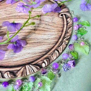 Op de afbeelding: Een bedelarmband met een zilverkleurige ketting versierd met paarse kralen, doorschijnende lichtgroene bladvormige bedels en kleine paarse bloemvormige kralen, uitgevoerd in diverse tinten violet en groen.