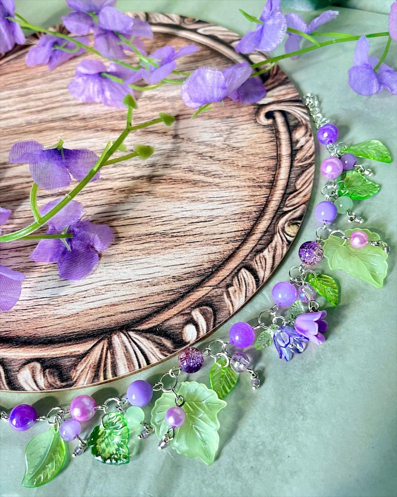 Op de afbeelding: Een bedelarmband met een zilverkleurige ketting versierd met paarse kralen, doorschijnende lichtgroene bladvormige bedels en kleine paarse bloemvormige kralen, uitgevoerd in diverse tinten violet en groen.