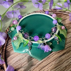 Op de afbeelding: Een zilverkleurige bedelarmband met paarse en lavendelkleurige kralen, doorschijnende groene bladvormige bedels en kleine paarse klokvormige bloemhangers. De armband is gemaakt van een metalen ketting en bevat diverse ronde kralen.