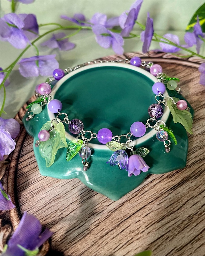 Op de afbeelding: Een zilverkleurige bedelarmband met paarse en lavendelkleurige kralen, doorschijnende groene bladvormige bedels en kleine paarse klokvormige bloemhangers. De armband is gemaakt van een metalen ketting en bevat diverse ronde kralen.