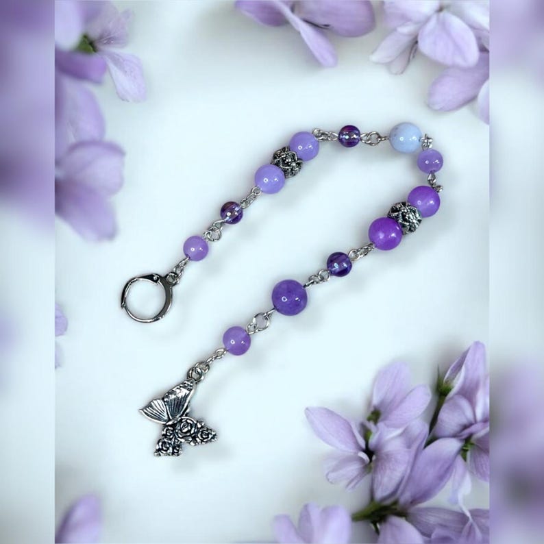 May include: A beaded keychain featuring a butterfly charm and a variety of purple beads. The beads range in color from light lavender to deep violet, connected by silver-toned metal links. The butterfly charm is detailed with floral patterns.