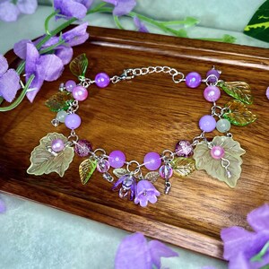 Op de afbeelding: Een zilverkleurige bedelarmband met paarse en lichtgroene kralen, doorschijnende groene bladvormige bedels en kleine paarse klokvormige bloemhangers. De armband is voorzien van een metalen ketting met een karabijnsluiting en een verlengketting.