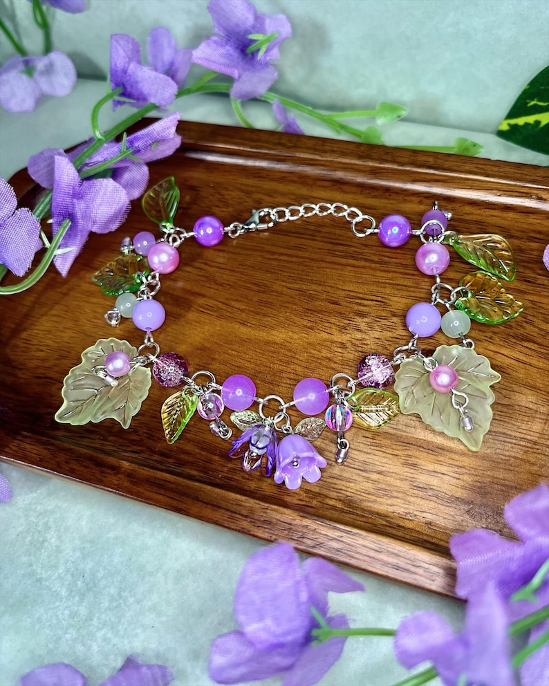 Op de afbeelding: Een zilverkleurige bedelarmband met paarse en lichtgroene kralen, doorschijnende groene bladvormige bedels en kleine paarse klokvormige bloemhangers. De armband is voorzien van een metalen ketting met een karabijnsluiting en een verlengketting.