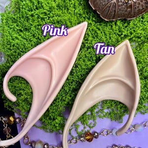 May include: Two pointed ear prosthetics, one in a light pink tone labeled Pink and one in a light tan tone labeled Tan. The ear pieces feature a curved design with a central ridge and a lower hook, presented as a pair of costume accessories.