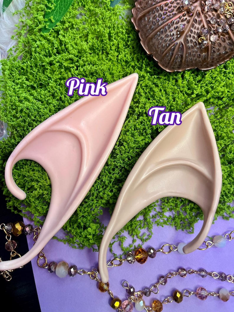May include: Two pointed ear prosthetics, one in a light pink tone labeled Pink and one in a light tan tone labeled Tan. The ear pieces feature a curved design with a central ridge and a lower hook, presented as a pair of costume accessories.