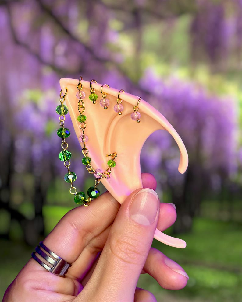 May include: A light-toned, pointed ear cuff accessory featuring a gold-tone chain adorned with faceted green and pink beads. The jewelry piece includes multiple small hoops attached to the upper edge and a longer chain draped across the ear structure.