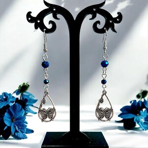 May include: Dangling earrings with a butterfly design. Each earring features a silver-toned teardrop pendant with a butterfly and a small flower. Dark blue beads are strung along the silver chain. The earrings are displayed on a black stand.
