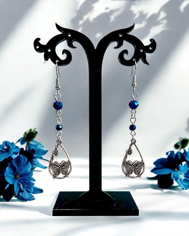 May include: Dangling earrings with a butterfly design. Each earring features a silver-toned teardrop pendant with a butterfly and a small flower. Dark blue beads are strung along the silver chain. The earrings are displayed on a black stand.