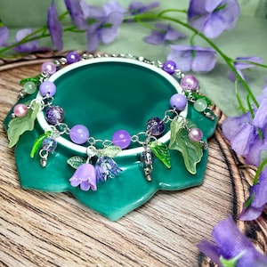 Op de afbeelding: Een zilverkleurige bedelarmband met paarse en lavendelkleurige kralen, doorschijnende groene bladvormige bedels en kleine klokvormige bloemhangers in tinten lichtpaars en blauw, verbonden door een fijn metalen schakelkettingontwerp.