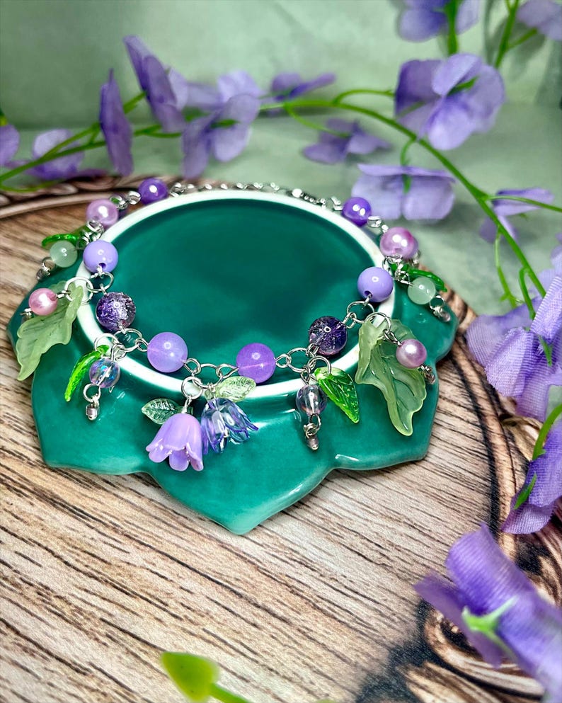 Op de afbeelding: Een zilverkleurige bedelarmband met paarse en lavendelkleurige kralen, doorschijnende groene bladvormige bedels en kleine klokvormige bloemhangers in tinten lichtpaars en blauw, verbonden door een fijn metalen schakelkettingontwerp.