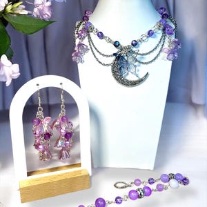 May include: A jewelry set featuring a necklace, earrings, and bracelet. The necklace has a silver crescent moon pendant with a butterfly, and purple beads. The earrings and bracelet also have purple beads and flower accents. The set is displayed on a white mannequin.
