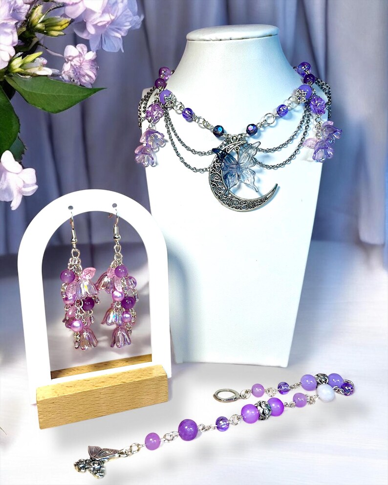 May include: A jewelry set featuring a necklace, earrings, and bracelet. The necklace has a silver crescent moon pendant with a butterfly, and purple beads. The earrings and bracelet also have purple beads and flower accents. The set is displayed on a white mannequin.