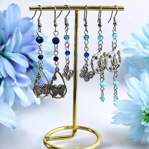 May include: A collection of silver-toned earrings displayed on a gold-colored earring stand. The earrings feature various designs, including butterfly pendants and beaded accents in shades of blue. The background includes blue flowers.