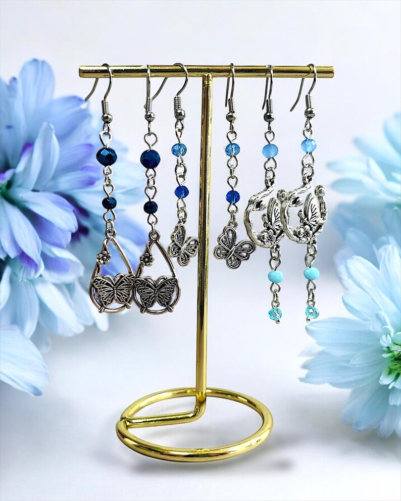 May include: A collection of silver-toned earrings displayed on a gold-colored earring stand. The earrings feature various designs, including butterfly pendants and beaded accents in shades of blue. The background includes blue flowers.