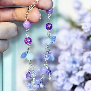May include: A delicate bracelet featuring a silver chain with dangling charms. The charms include iridescent purple and lavender beads, small butterfly and flower shapes, and teardrop-shaped accents. The bracelet is held up against a soft purple floral background.