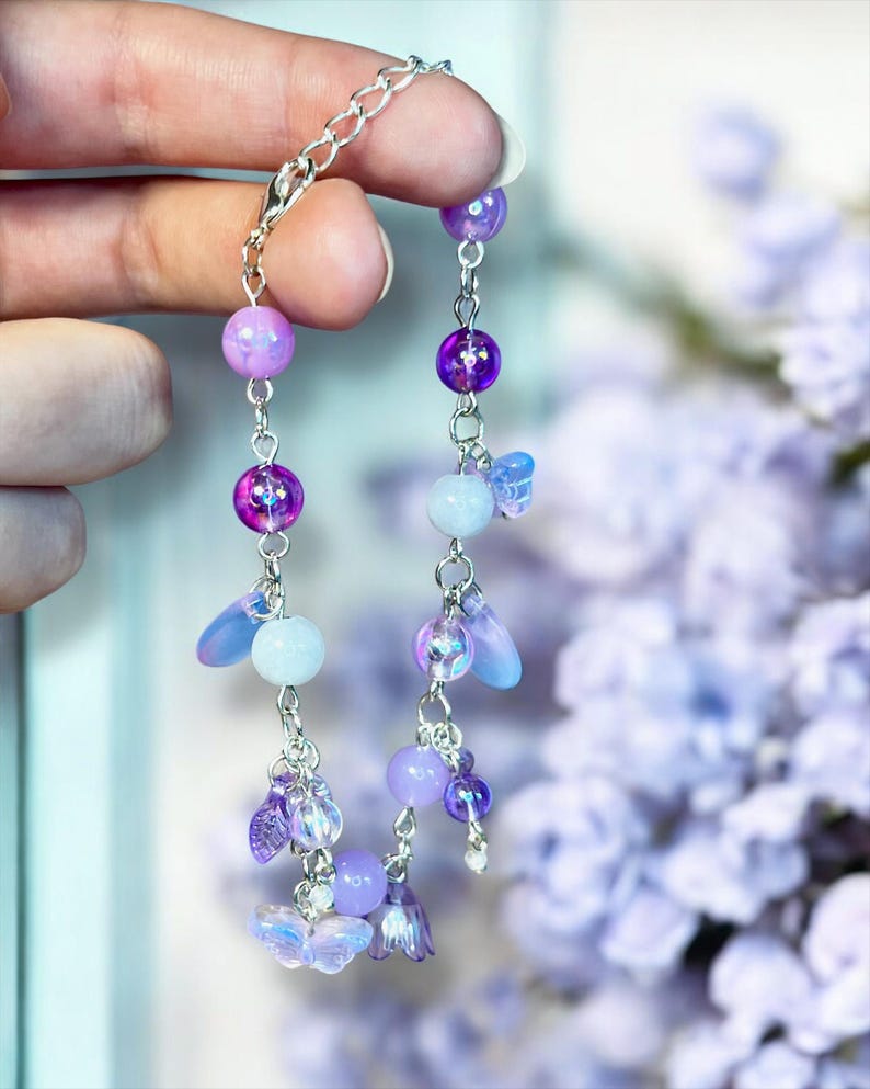 May include: A delicate bracelet featuring a silver chain with dangling charms. The charms include iridescent purple and lavender beads, small butterfly and flower shapes, and teardrop-shaped accents. The bracelet is held up against a soft purple floral background.